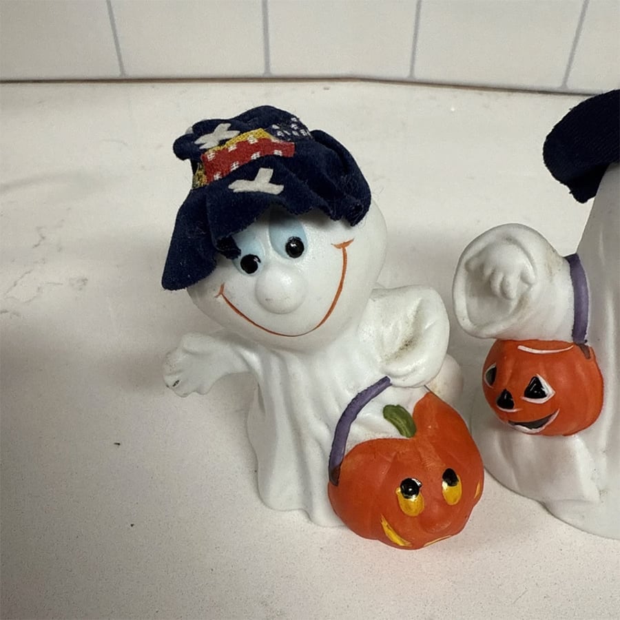 A smiling ghost figurine with a blue hat holds an orange pumpkin bucket. Another partially visible ghost with a similar pumpkin bucket stands beside it on a white surface.