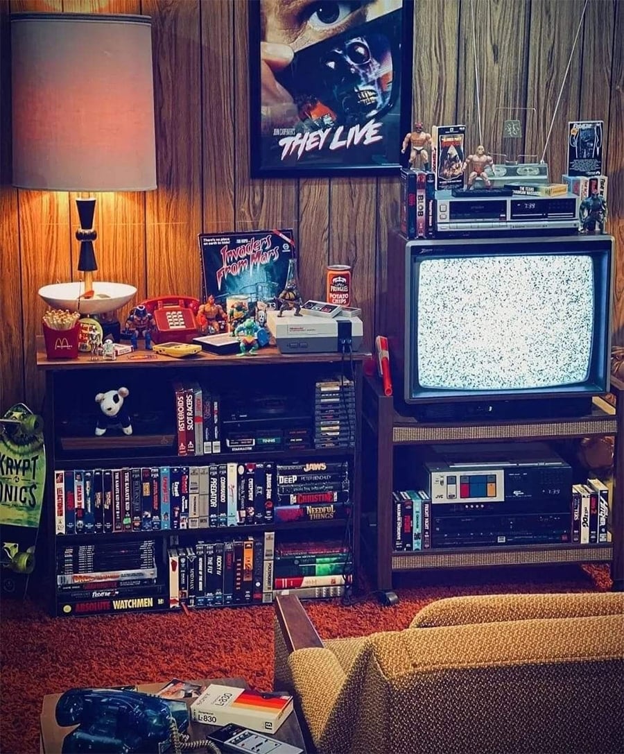 A retro living room with a CRT TV showing static, VHS tapes, video game consoles, action figures, and a board game on shelves. A "They Live" poster hangs on a wood-paneled wall.