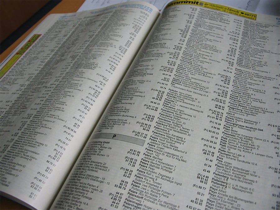 An open phone book showing columns of names, addresses, and phone numbers printed in black on white pages. The book is spread out on a desk or table.