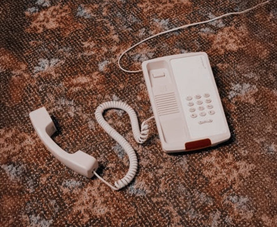 A beige corded landline telephone with the receiver off the hook rests on a patterned carpeted floor.