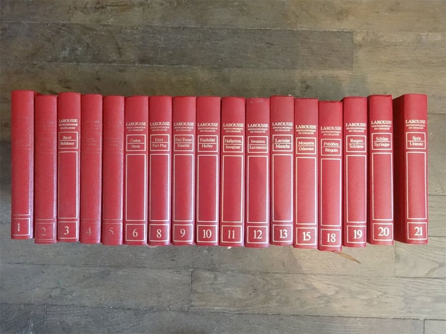 A set of 21 red hardcover encyclopedias with gold lettering is arranged in order on a wooden floor. Each book is labeled with a volume number and topic on the spine.