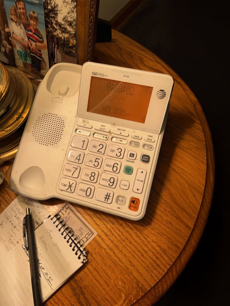 A white AT&T landline phone with large buttons sits on a wooden table next to a notepad with handwritten notes and a pen. Framed family photos and a lamp are partially visible in the background.
