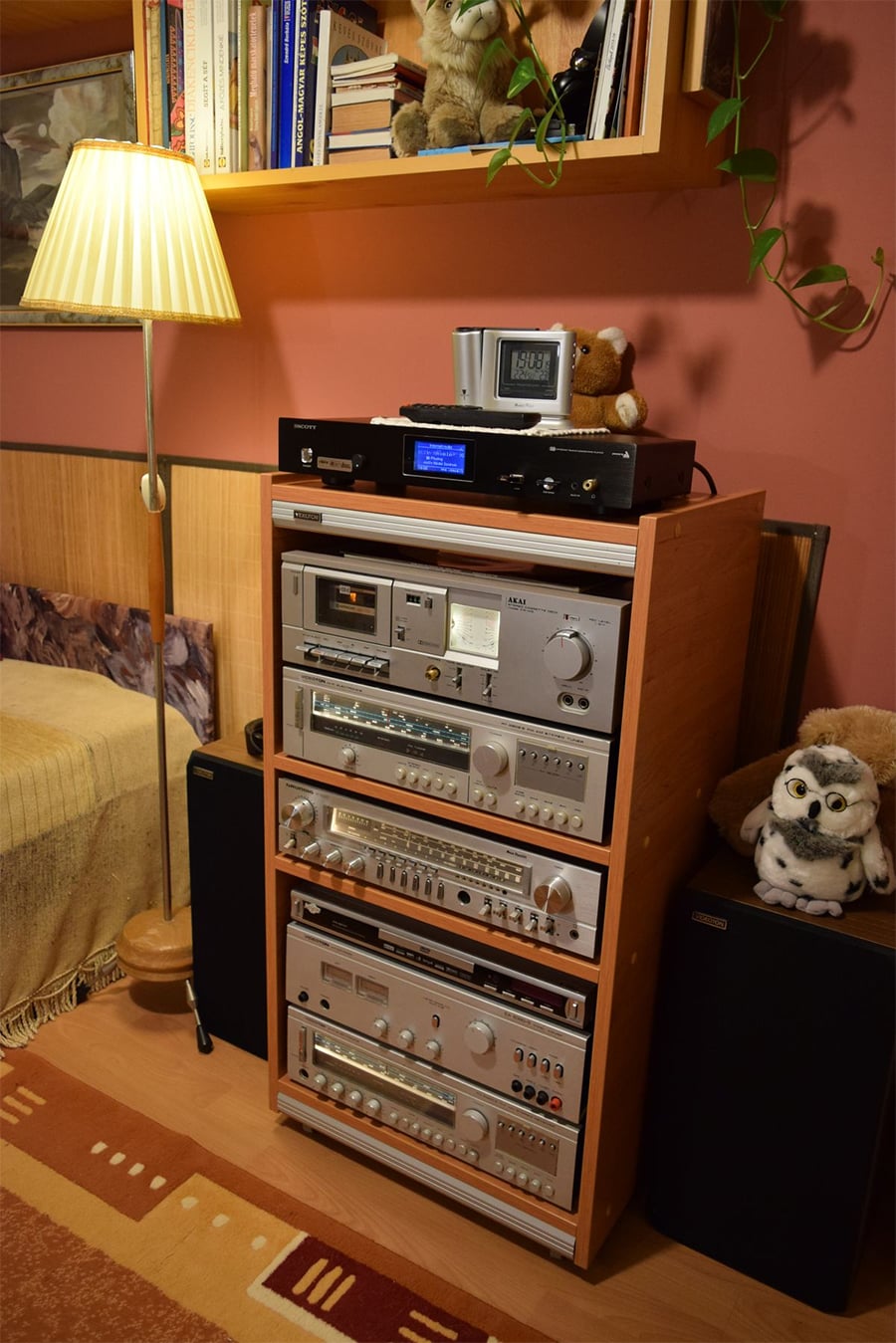A wooden stereo rack stands against a salmon wall, filled with vintage audio components, including an Akai cassette deck and several tuners. A tall floor lamp, books on a shelf, and plush owl toys decorate the cozy, retro-style room.