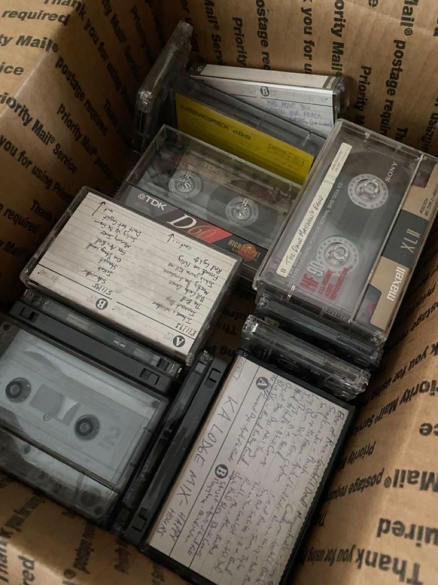 A cardboard box filled with several cassette tapes in plastic cases, some with handwritten labels and song lists visible on the front of the cases.