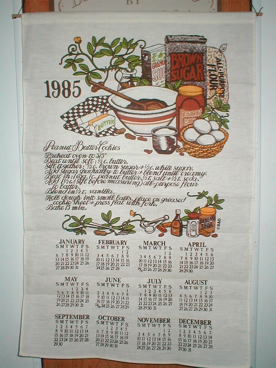 1985 calendar tea towel with a peanut butter cookie recipe and illustrated baking ingredients, including eggs, butter, flour, brown sugar, and utensils. Each month is listed at the bottom in a grid format.