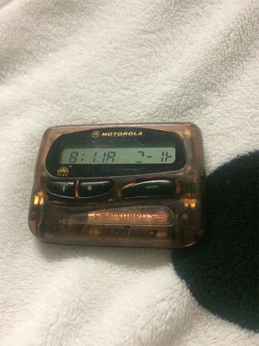 A brown Motorola pager displays "8:11A 7-11" on its screen, resting on a white textured fabric with a black spot.