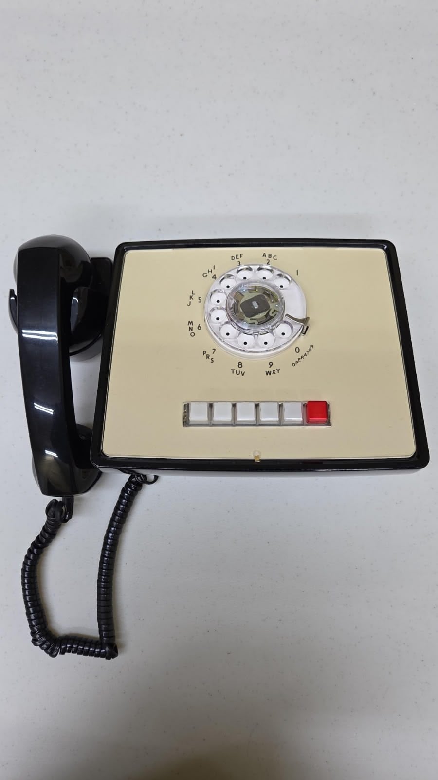 A vintage rotary dial telephone with a beige faceplate, black handset, coiled cord, and five rectangular buttons, one of which is red, placed on a light grey surface.