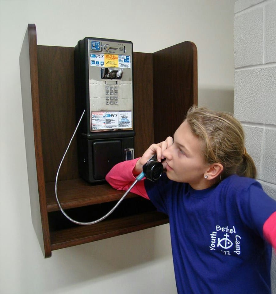A girl in a blue Youth Bethel Camp T-shirt is using a black payphone mounted on a wooden wall shelf inside a building.
