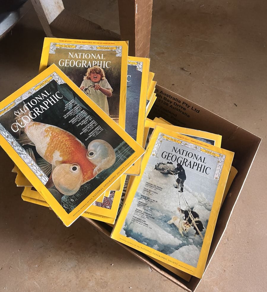 A box filled with old National Geographic magazines, each with the iconic yellow border. The visible covers feature images of a cephalopod, a polar explorer, and a person with a catch. The box sits on a tan floor.