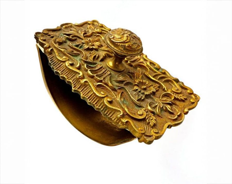 Ornate vintage brass blotter with floral and scroll designs on the top, featuring a rounded handle and a curved base, photographed against a plain white background.