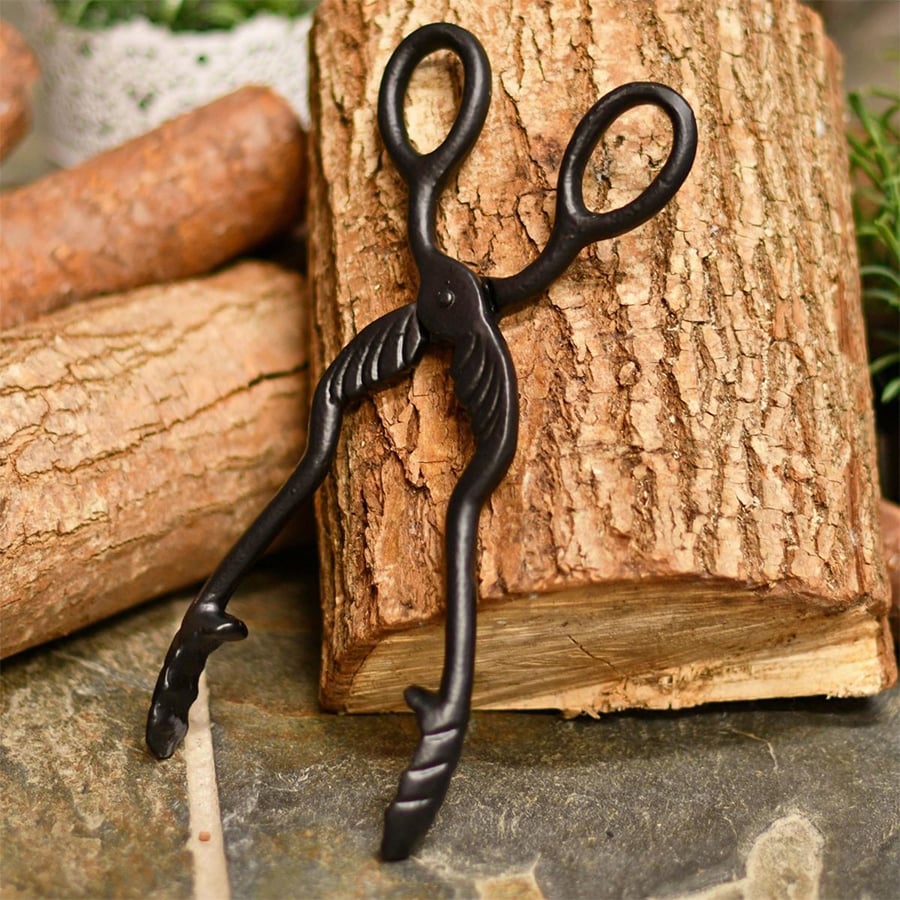 Black metal tongs with decorative wing-like details rest on a cut log, surrounded by other wooden logs and greenery.