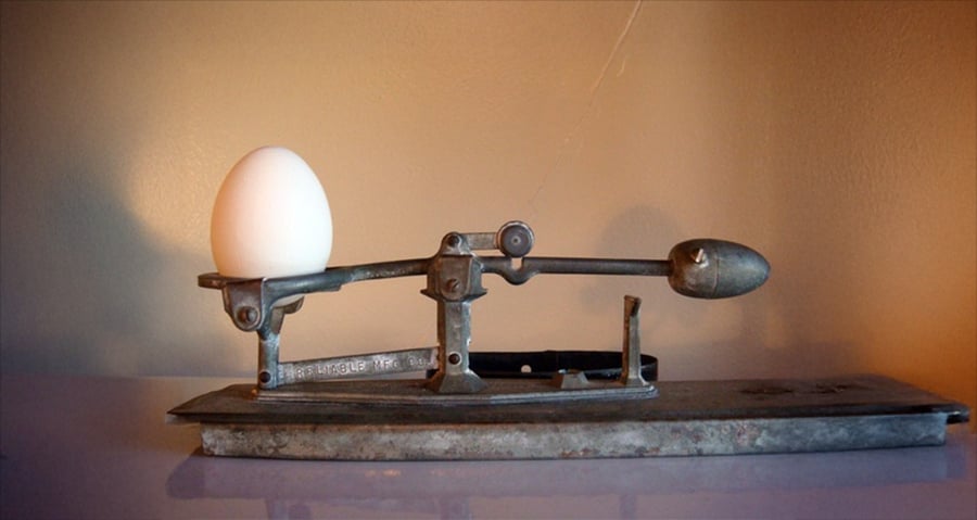 A single white egg sits on one side of an old-fashioned metal balance scale against a neutral background.