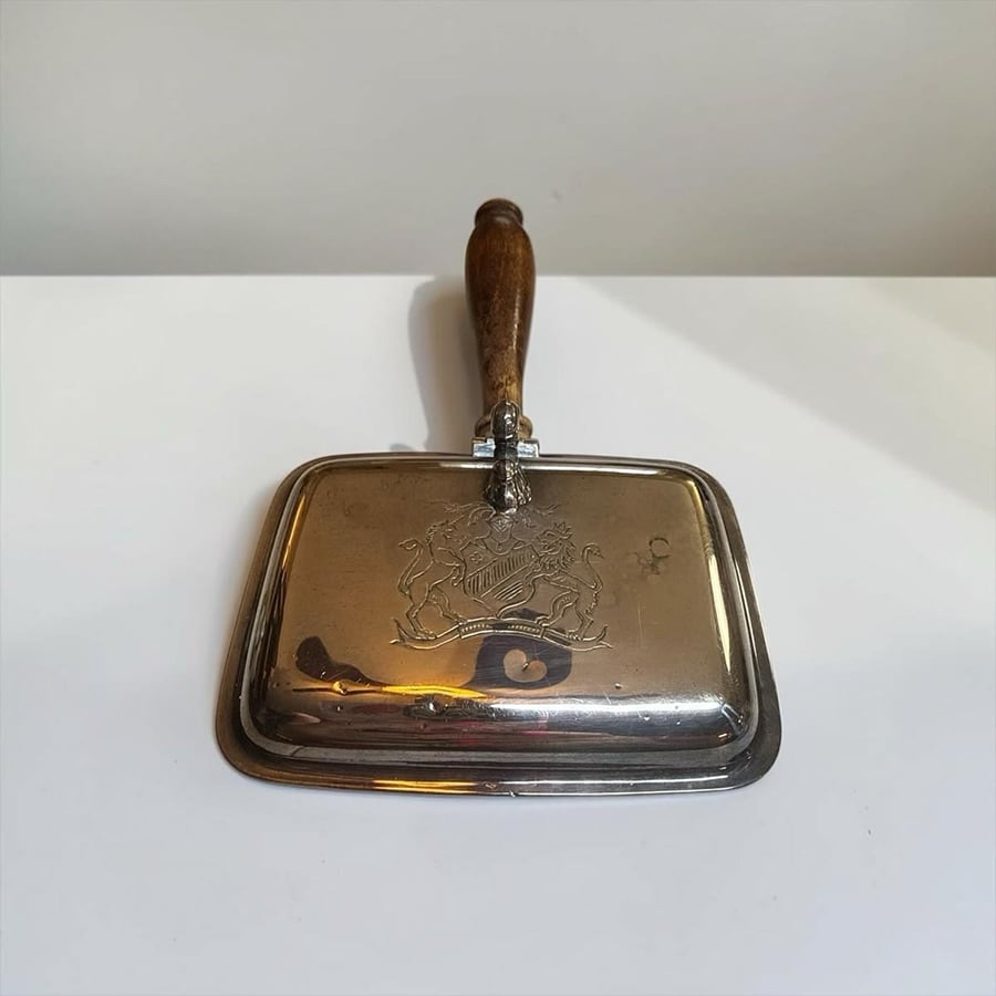 A vintage metal crumb catcher with a wooden handle sits on a white surface. The metal pan has an engraved design and slight reflective marks.