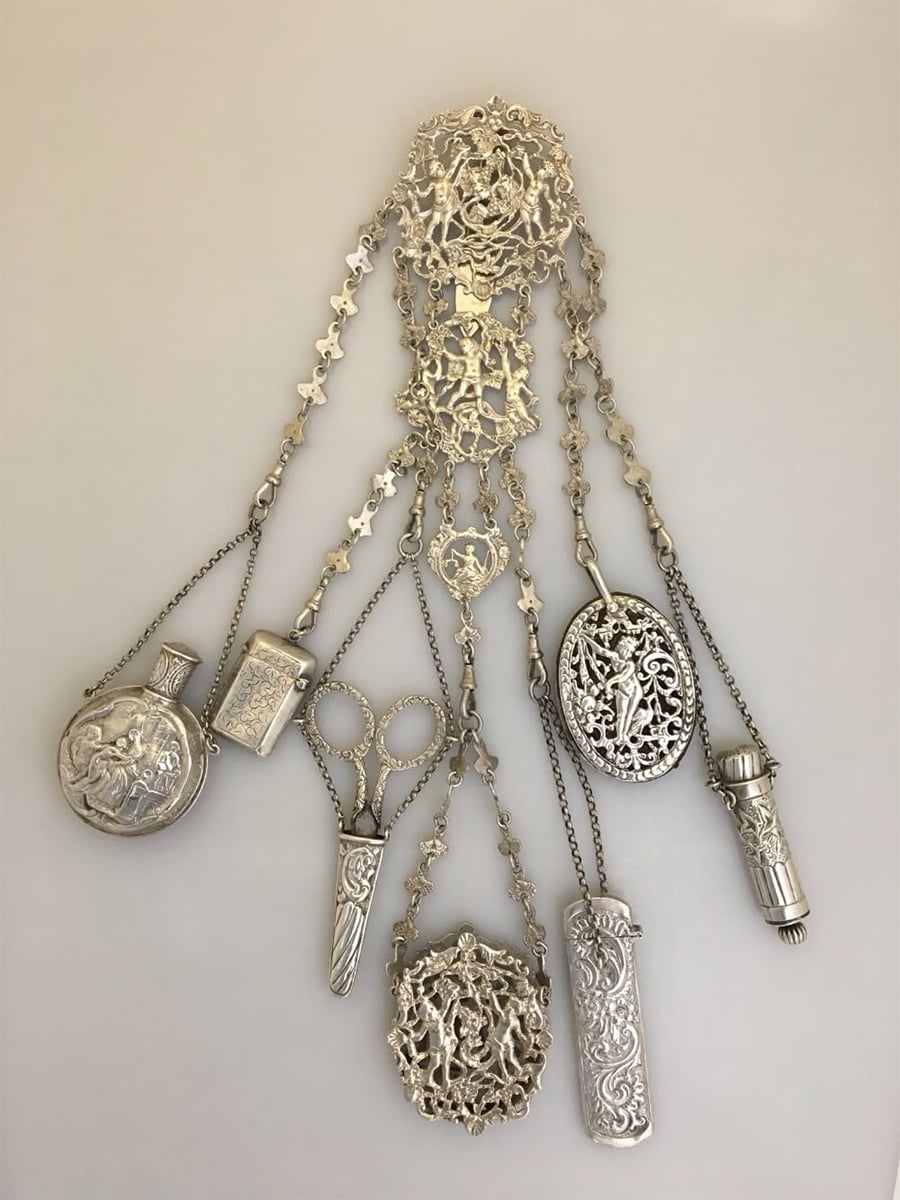 A vintage silver chatelaine with multiple ornate chains holds small items like scissors, a pill box, and other decorative accessories, all crafted in intricate silver designs, displayed on a white background.