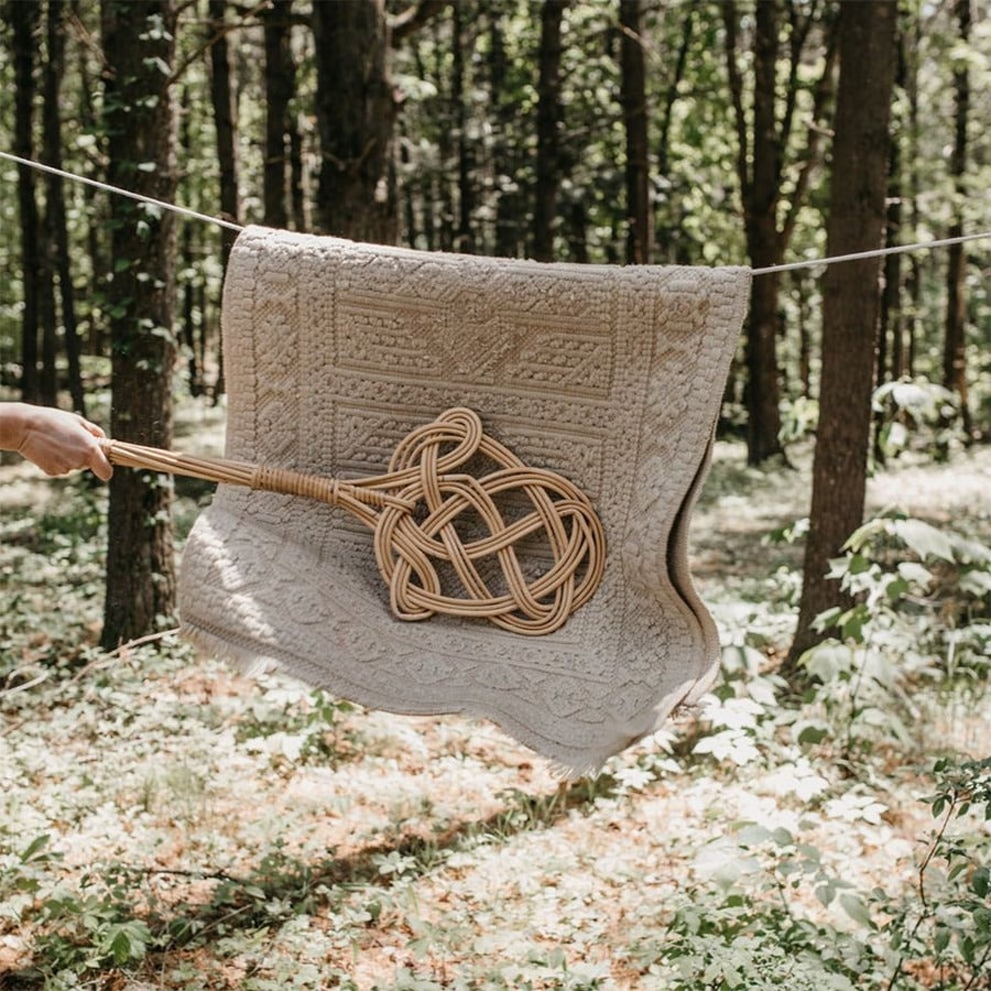 A hand holds a woven carpet beater, hitting a patterned beige rug hanging on a clothesline in a sunlit forest clearing.