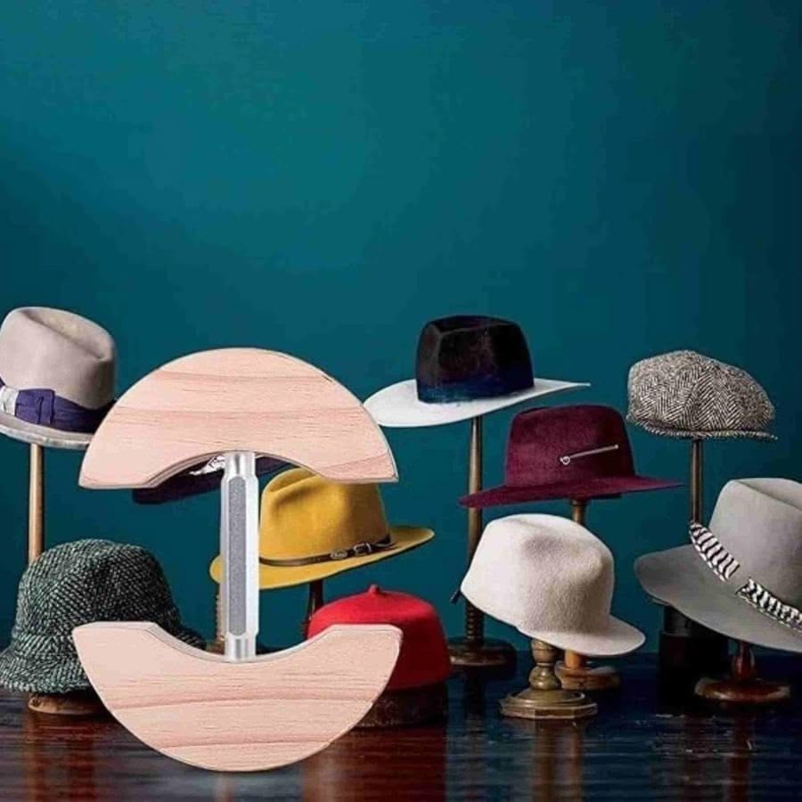 Several stylish hats on stands are displayed on a wooden surface against a blue wall. In the foreground, there’s an unusual wooden clamp-like device with semicircular ends.