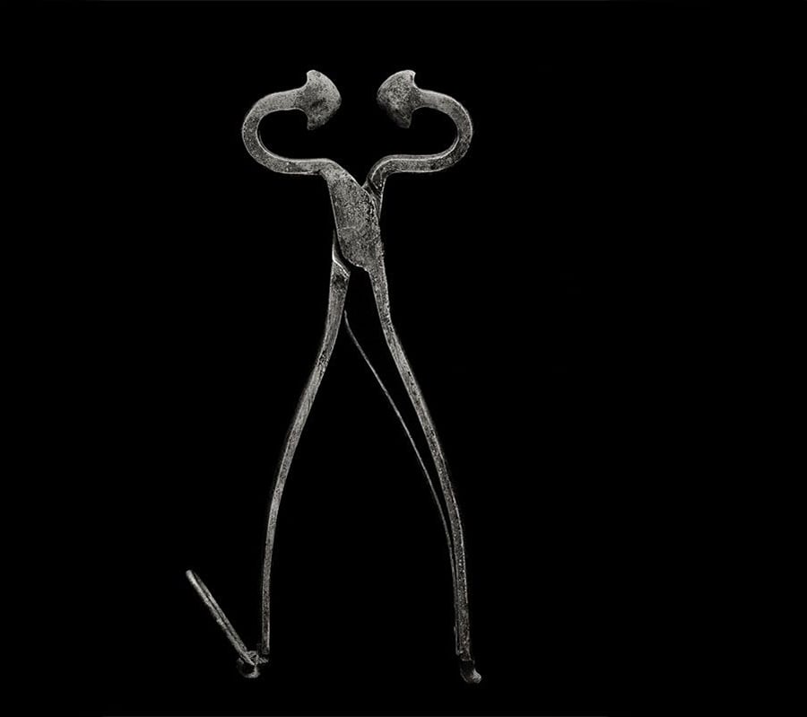 Ancient metal tool with looped handles and long, curved prongs, resembling tongs or pincers, displayed on a black background.