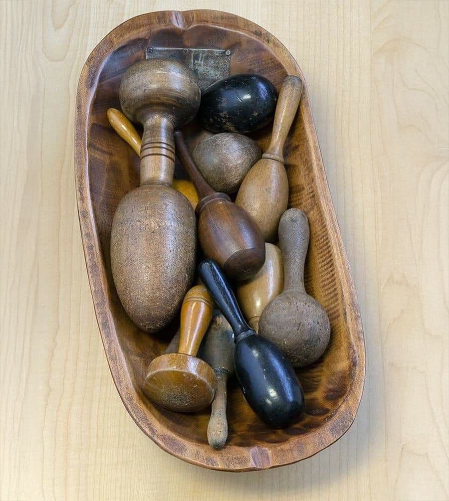 A wooden bowl filled with various vintage wooden sock darners and fabric stretchers, in different shapes, sizes, and finishes, sits on a light wood surface.