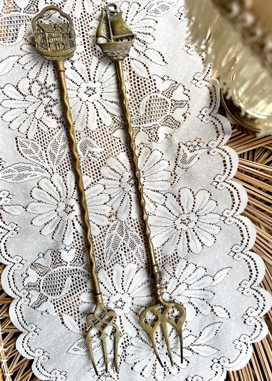 Two ornate brass cocktail forks with decorative handles—one featuring a building, the other a sailboat—rest on a white lace doily atop a wicker surface, with a glass partly visible on the right.
