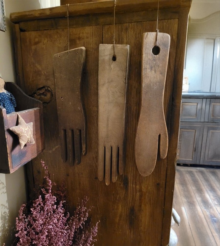 Three wooden glove forms with finger shapes hang by strings on a rustic wooden cabinet. Dried purple flowers sit below, and a box with fabric stars decorates the left side. A kitchen is visible in the background.