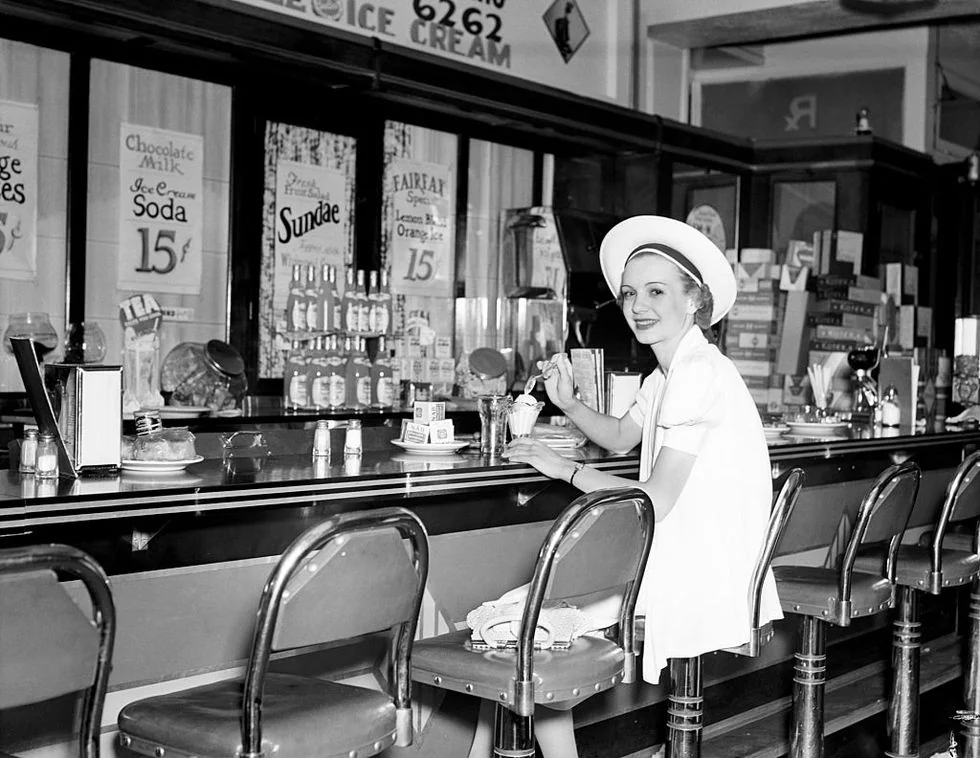 A woman in a white uniform sits at a vintage soda fountain counter, smiling and holding a drink. Behind her are shelves with jars and signs advertising sundaes, sodas, and ice cream.
