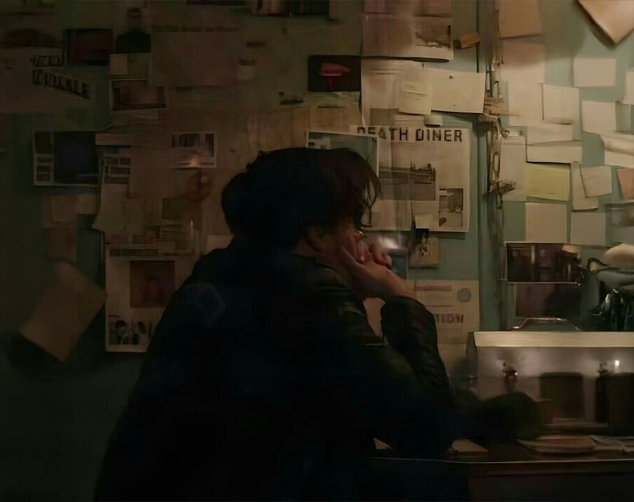 A person sits at a cluttered desk in a dimly lit room, facing a wall covered with notes, papers, and photos. The scene has a mysterious, investigative atmosphere.