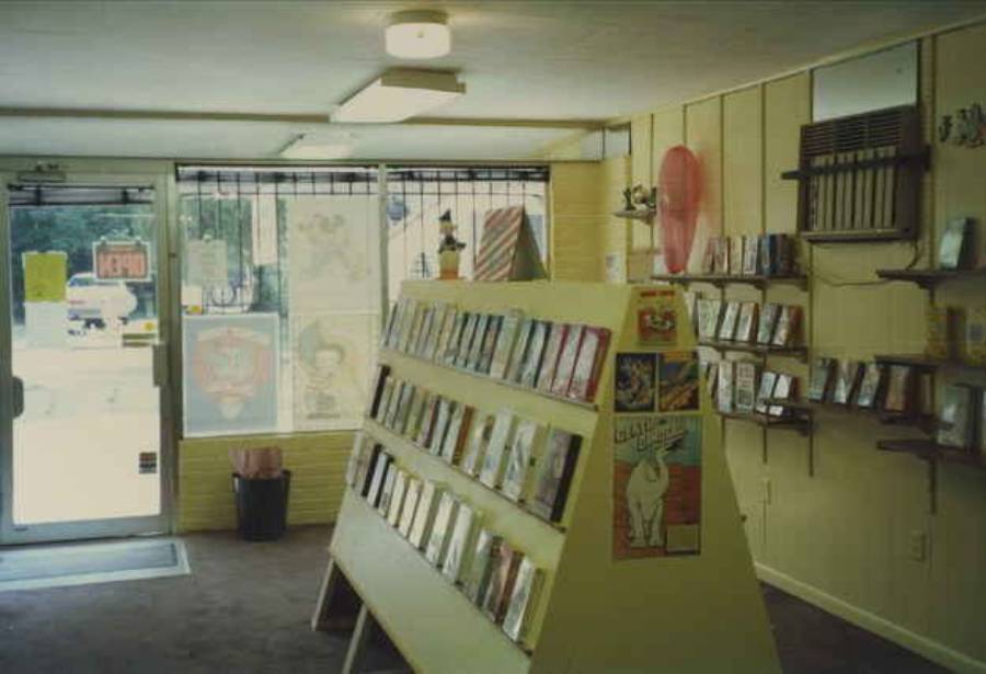 A small, vintage video store with rows of VHS tapes, a display rack in the center, shelves on the wall, and a window with posters. An open sign is on the glass door.