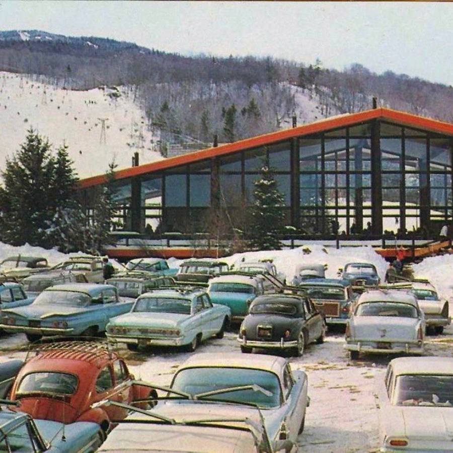 A snowy ski resort parking lot with vintage cars parked in front of an A-frame lodge. Snow-covered mountains and trees are visible in the background.