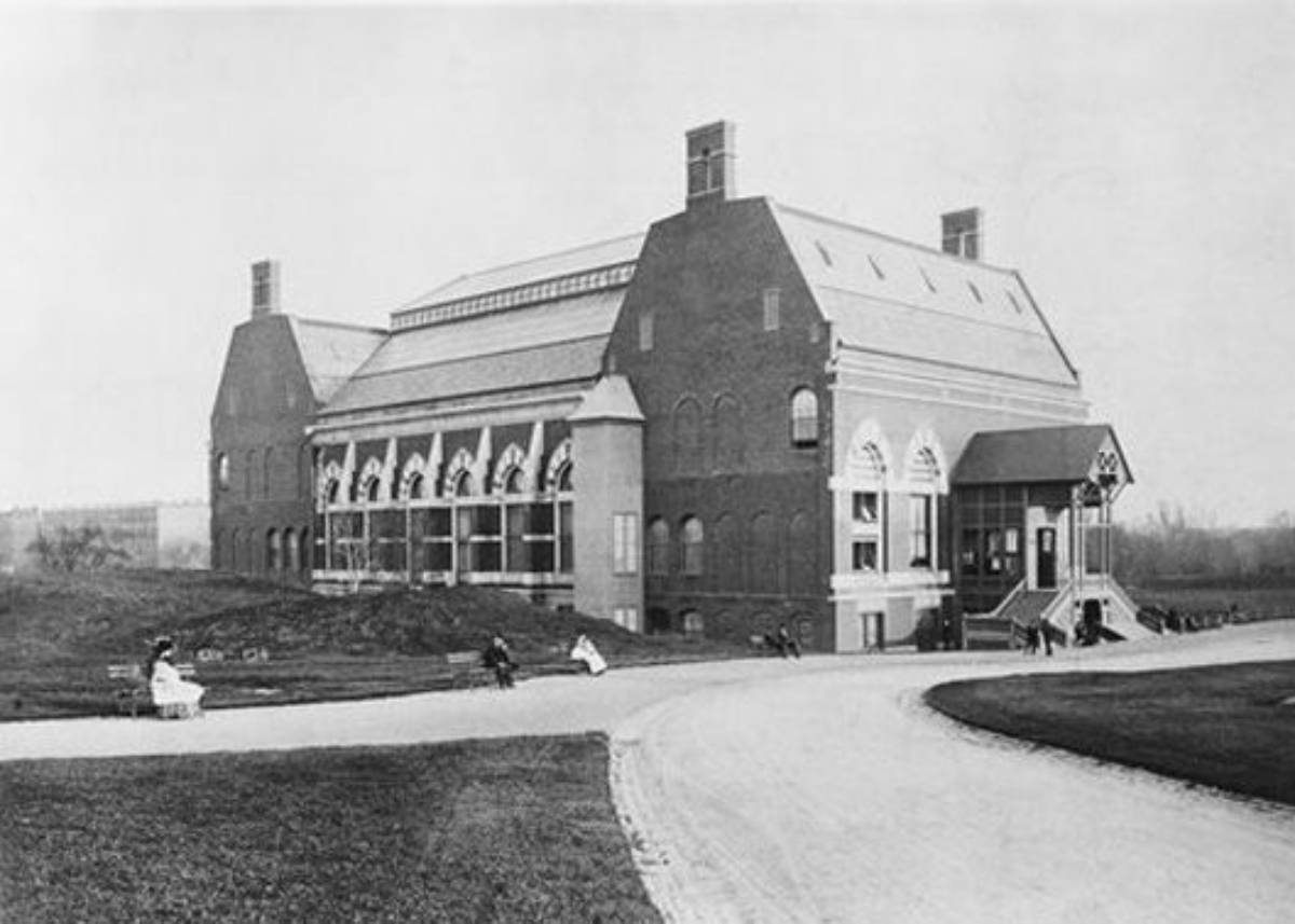 A large brick building with arched windows sits beside a winding path. Several people in old-fashioned clothing are visible on the grass and pathway in front of the building. The scene appears historical.