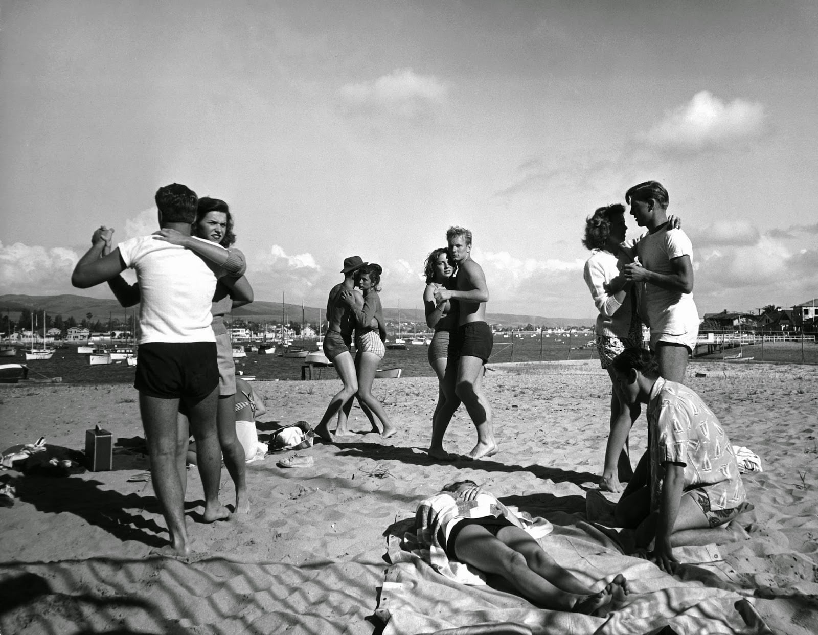 A group of young adults dance and relax on a sandy beach, some standing and some sitting on towels. There are boats and buildings in the background under a partly cloudy sky.