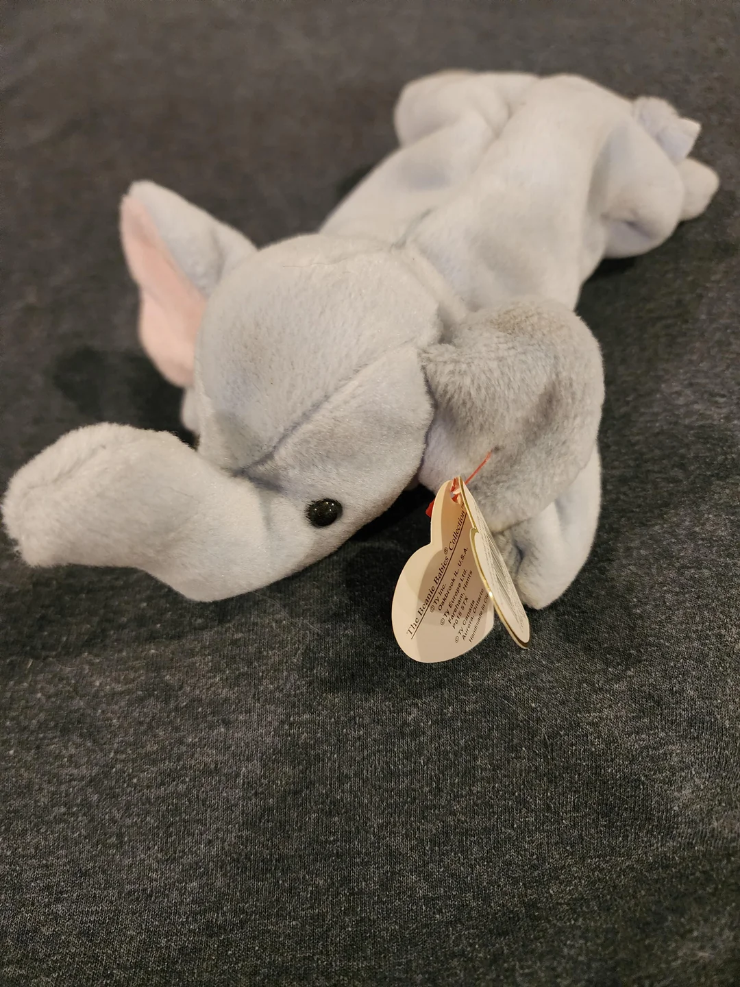 A gray plush elephant toy with a curved trunk and pink inner ears rests on a dark surface. A tag is attached to its ear.