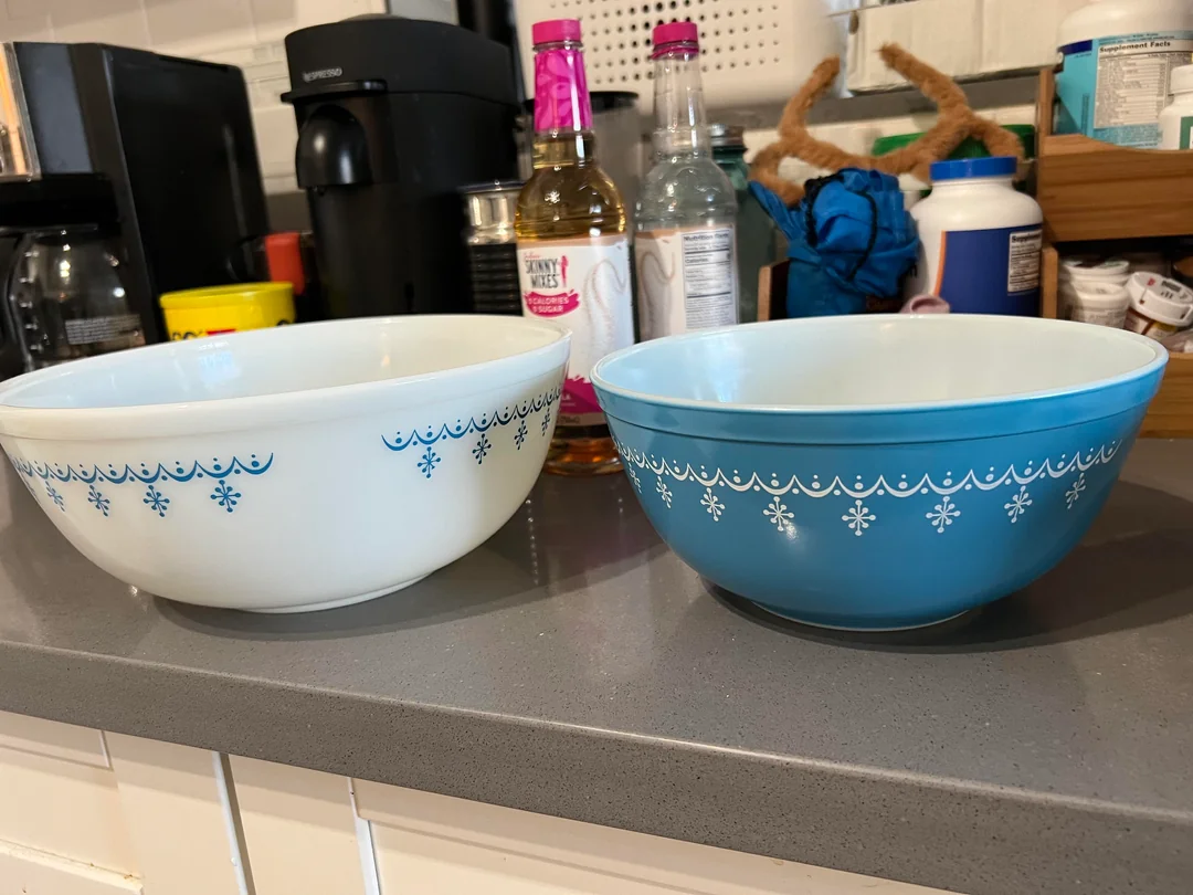 Two vintage mixing bowls sit on a kitchen counter; one is white with a blue pattern, and the other is blue with a white pattern. Various kitchen items are in the background.