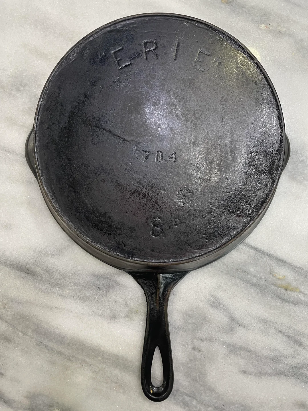 A vintage black cast iron skillet marked “ERIE” sits on a marble surface. The pan also displays the numbers “704” and “8” along with a small symbol, indicating its size and model.