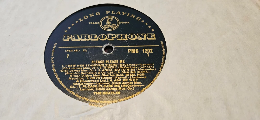 A close-up of a vintage Parlophone vinyl record label for “Please Please Me” by The Beatles, showing track names and credits in gold text on a black background.