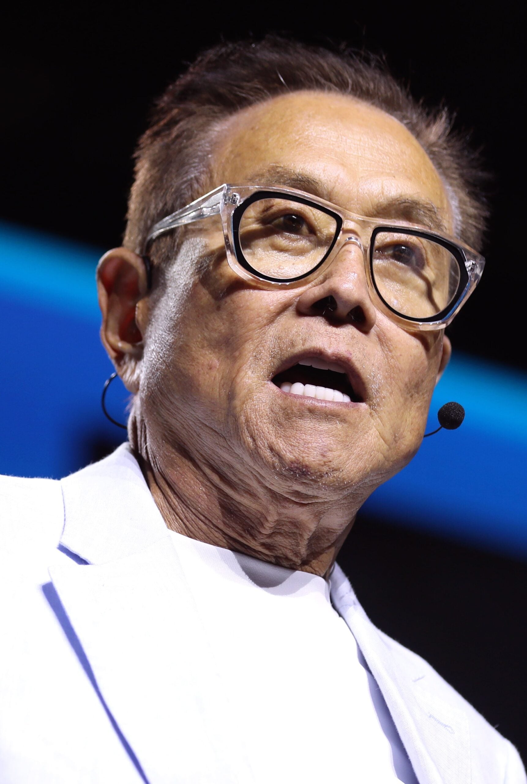A man wearing glasses and a headset microphone speaks on stage, dressed in a light-colored jacket and white shirt, with a dark background behind him.