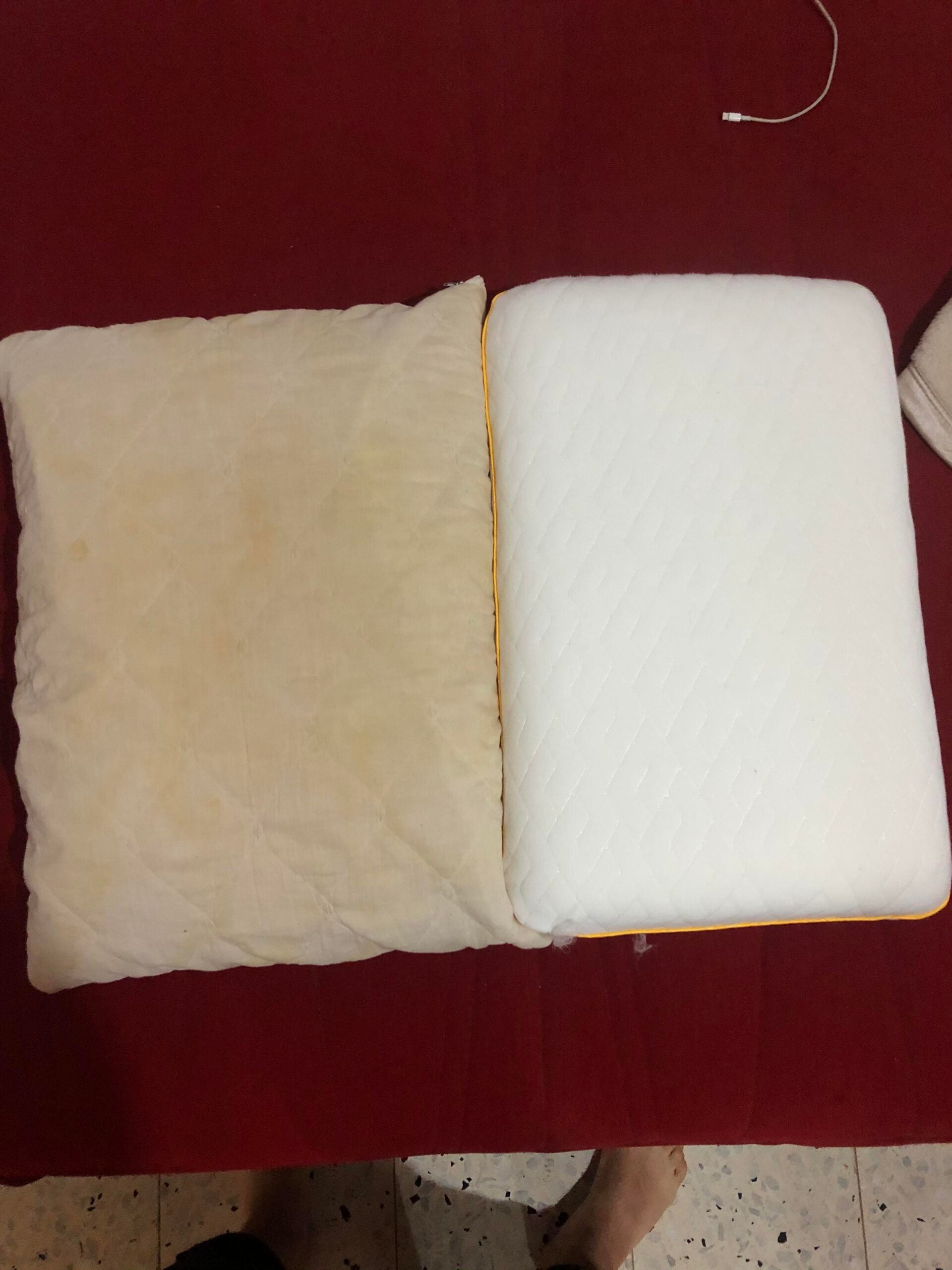 Two pillows on a red surface: the left one is yellowed and stained, while the right one is bright white with a textured cover. A charging cable is visible in the top right corner.