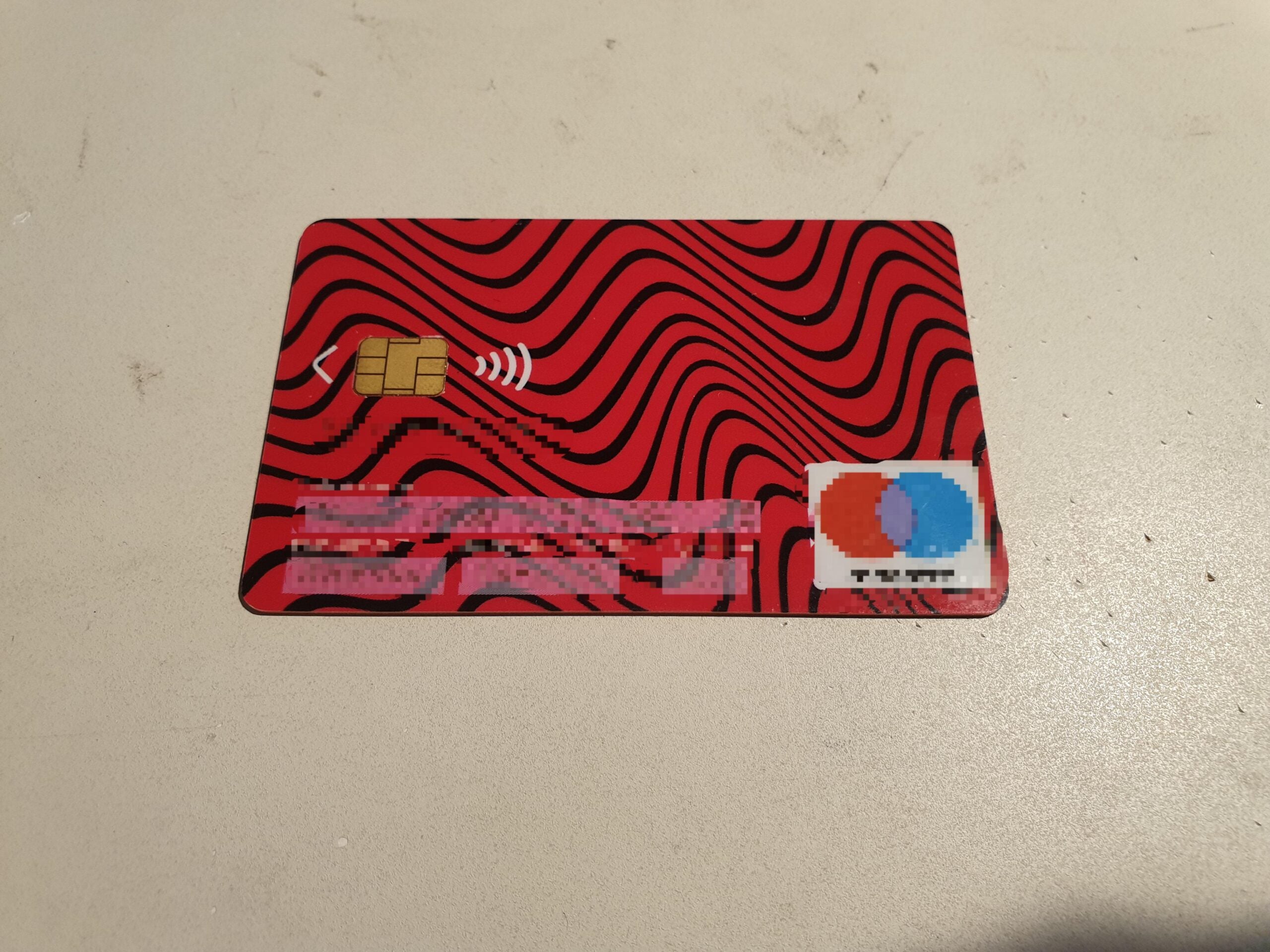 A red debit or credit card with wavy black lines, a chip, and contactless symbol sits on a light surface. Card details and logo are pixelated for privacy.