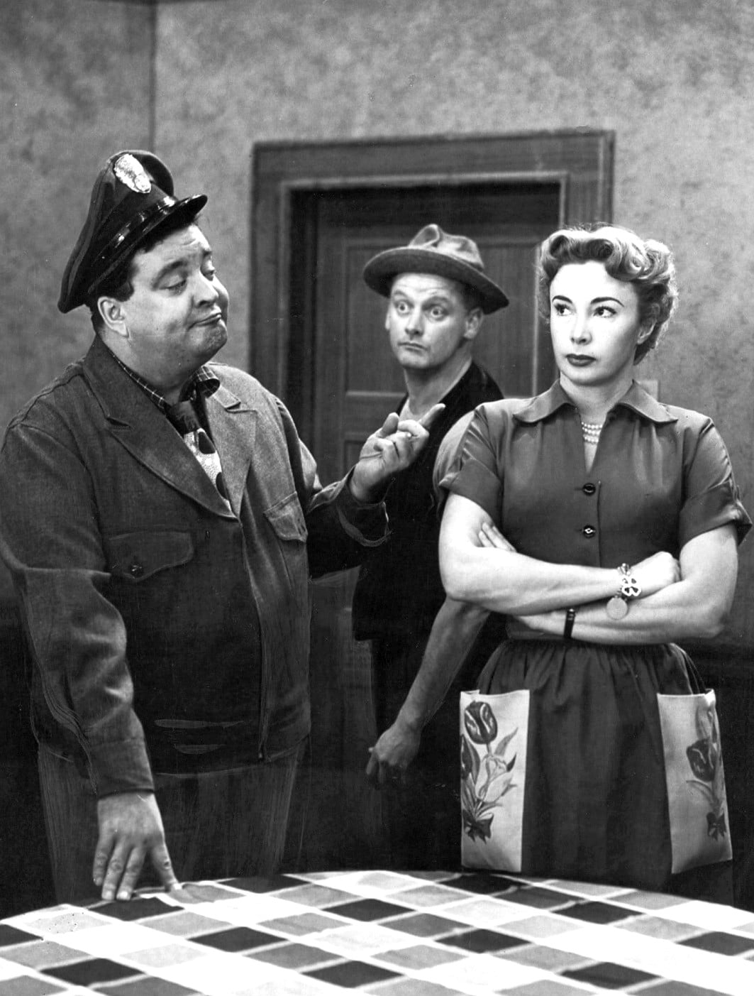 A man in a uniform gestures while talking to a woman with crossed arms. Another man in a cap stands behind them, looking surprised. They are indoors, next to a checkered table.