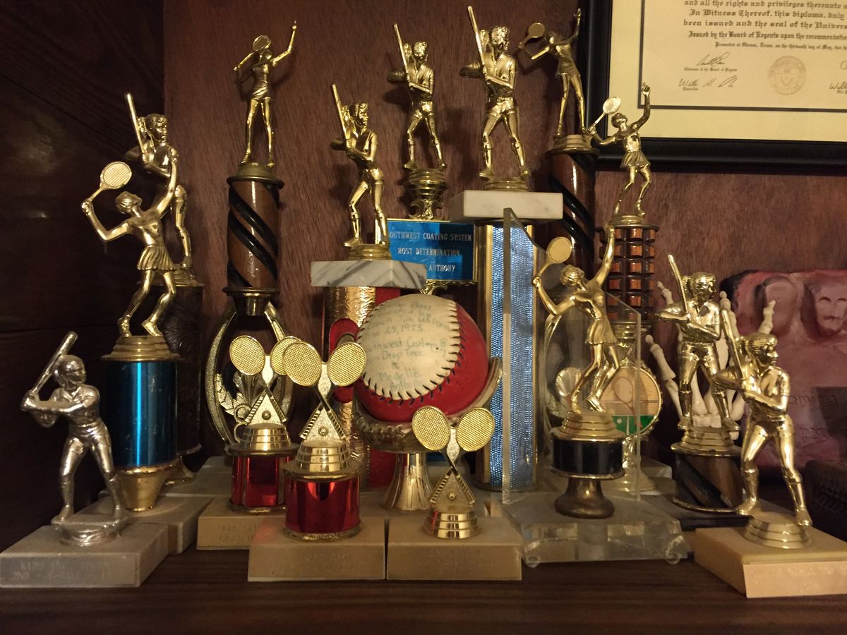 A collection of sports trophies, mainly for baseball and tennis, arranged on a wooden shelf, with a signed baseball and certificate in the background.