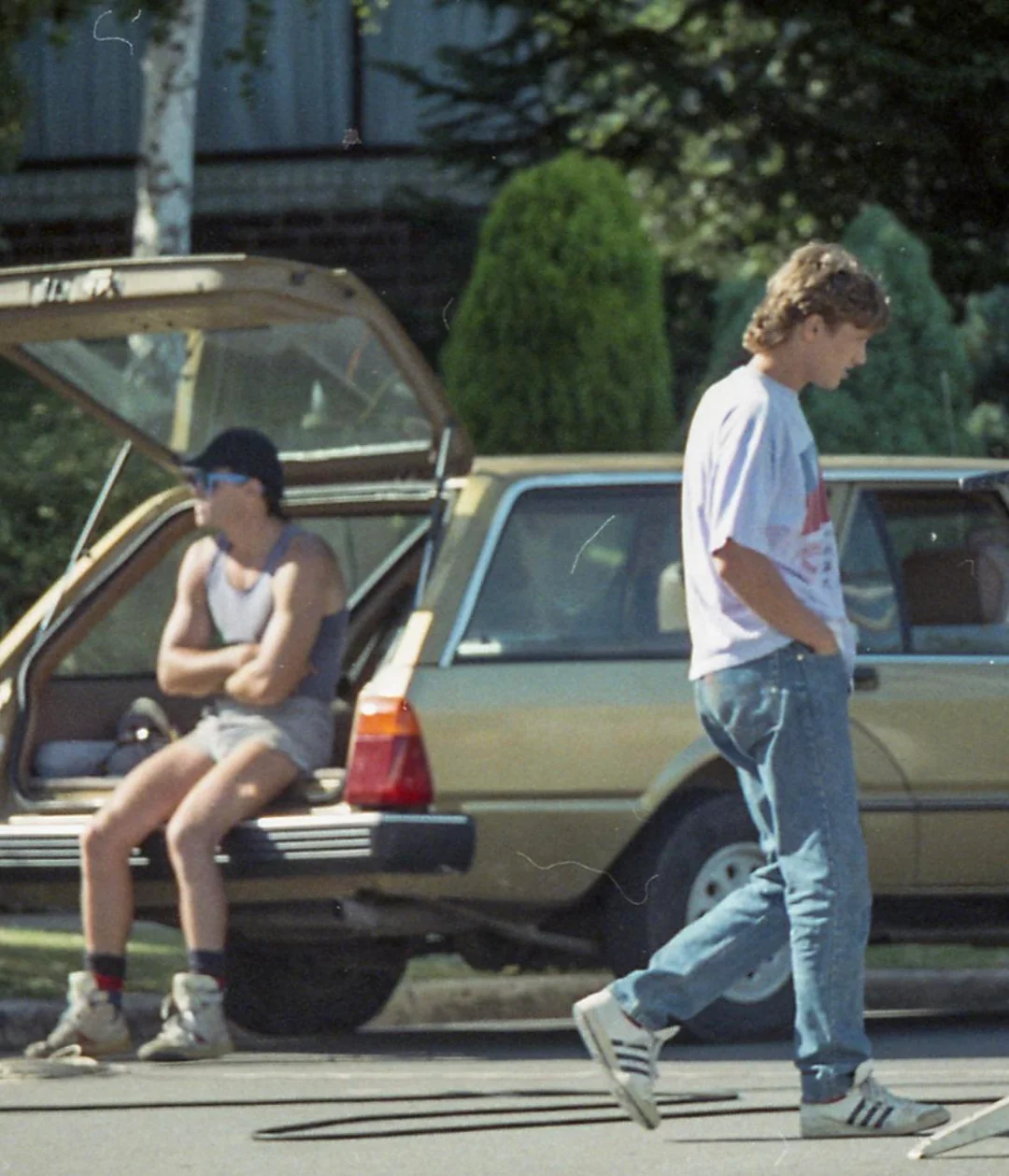Two people near a parked car: one sits in the open trunk wearing shorts, a tank top, and a cap, while the other walks by in jeans and a t-shirt. Trees and greenery are in the background.