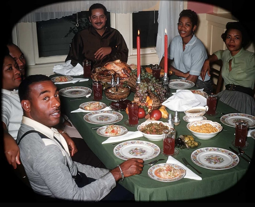 Six people sit around a table set with food, drinks, candles, and decorative fruit, smiling and enjoying a meal together in a warmly lit dining room.
