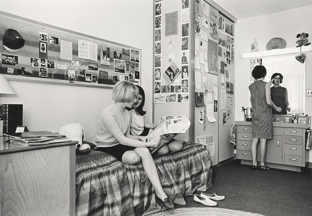 Two women sit on a bed looking at a newspaper in a dorm room filled with photos and notes. Another woman stands at the sink, looking in the mirror. Shoes are on the floor near the bed.