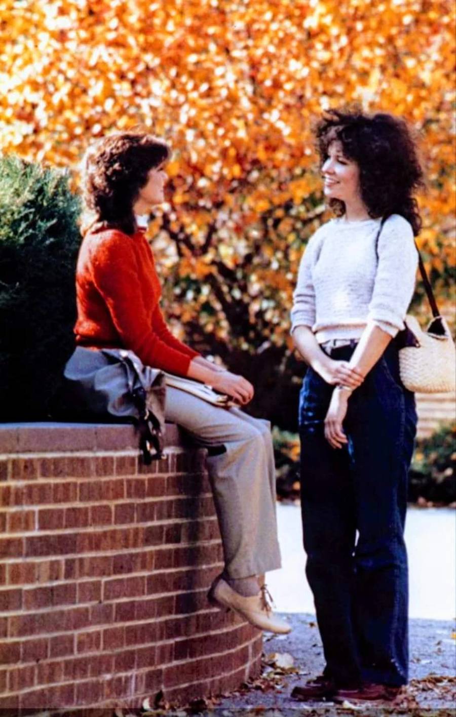Two women talk outside by a brick wall, with autumn leaves in the background. One is sitting with a red sweater and gray pants, the other is standing, wearing a white sweater and jeans, holding a straw bag.