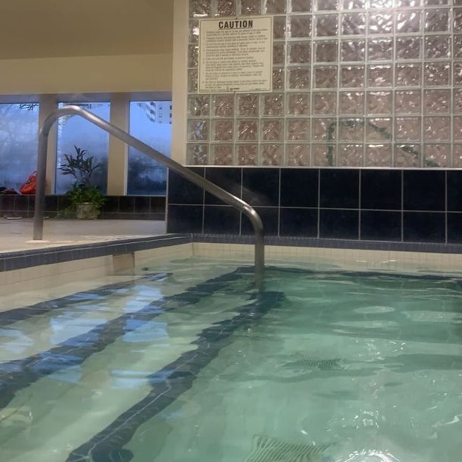 Indoor swimming pool with clear water, metal handrail, blue and white tiled edge, and a caution sign on a glass block wall. Some plants and lounge chairs are visible in the background.