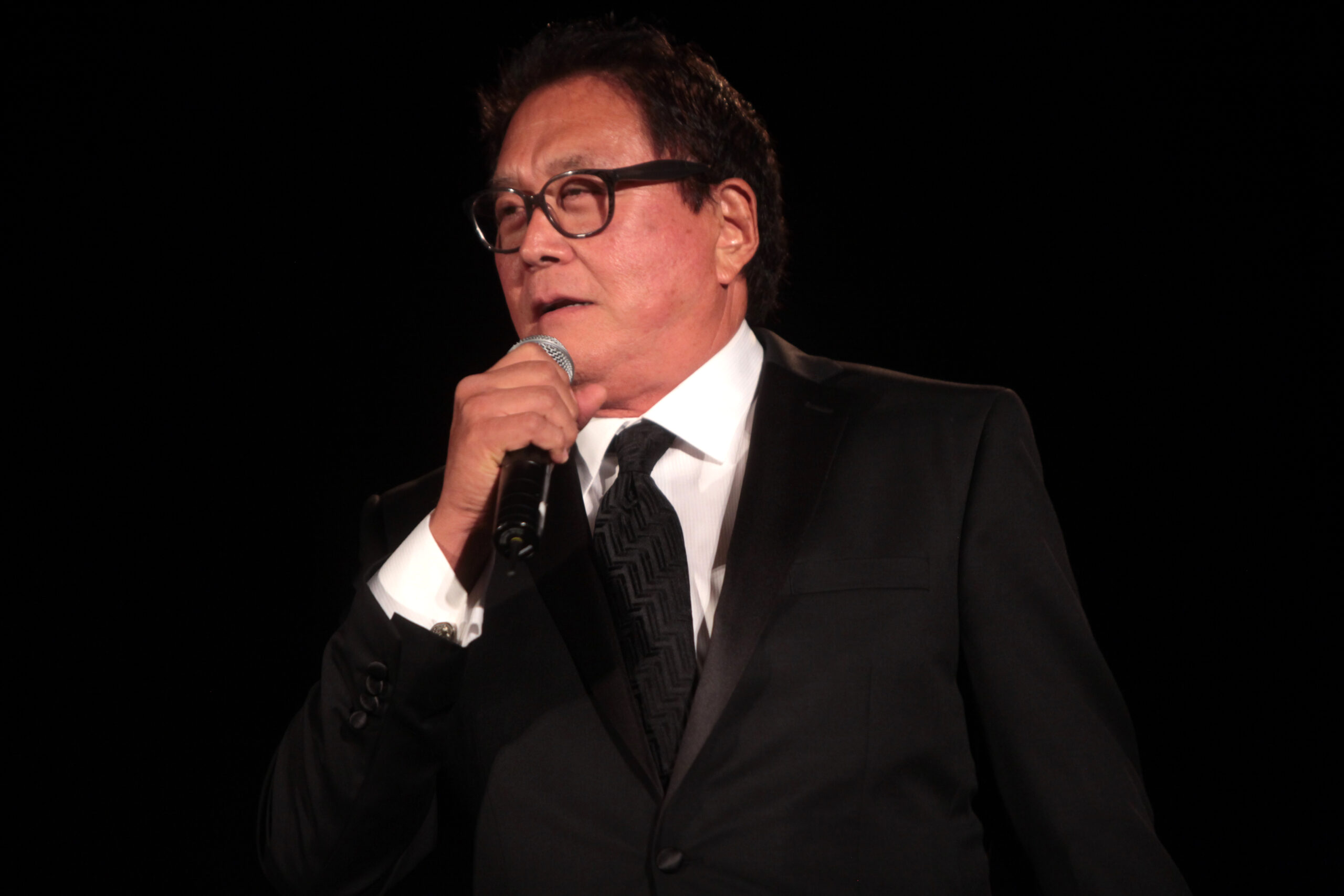 A man wearing glasses and a black suit holds a microphone and speaks on stage against a dark background.