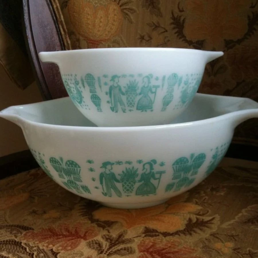 Two white mixing bowls with teal blue vintage folk art designs, featuring people, flowers, and roosters, sit stacked on an ornate upholstered chair with a floral pattern.