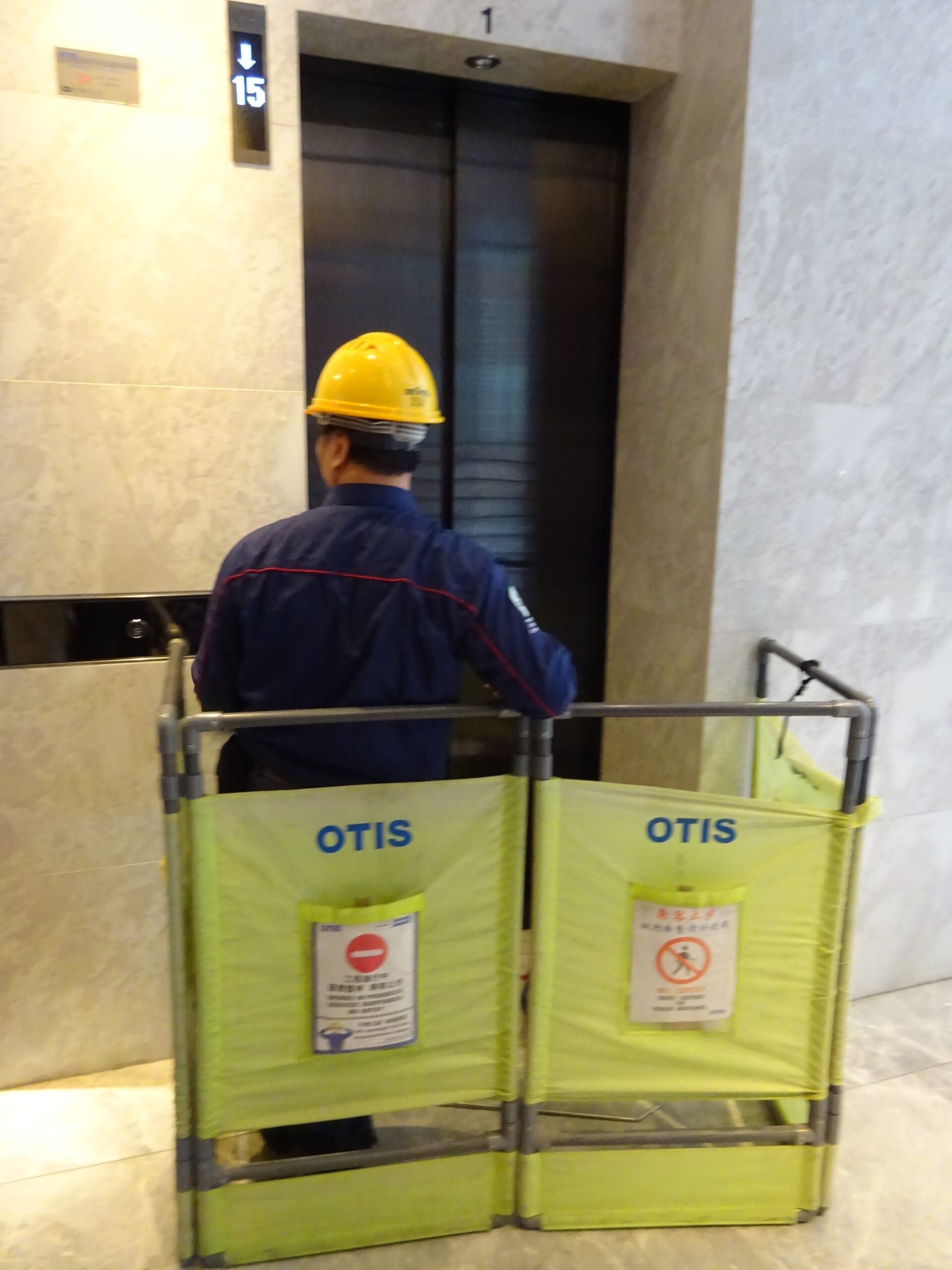 A worker in a yellow hard hat stands in front of a closed elevator, behind a yellow safety barrier labeled “OTIS” with warning signs, in a building lobby with marble walls.