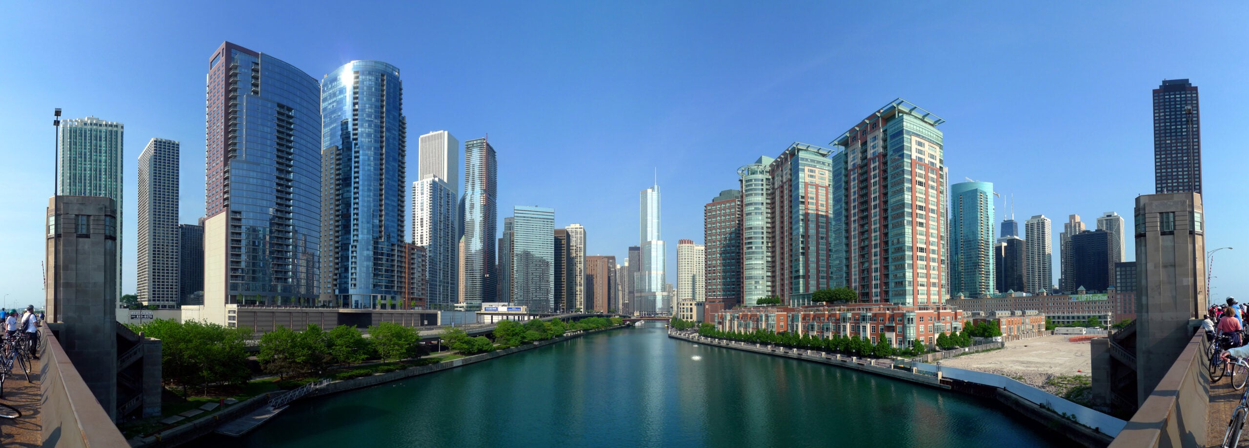A wide view of a modern city skyline with tall glass buildings reflected in a river, under a clear blue sky, with greenery and people along the waterfront.