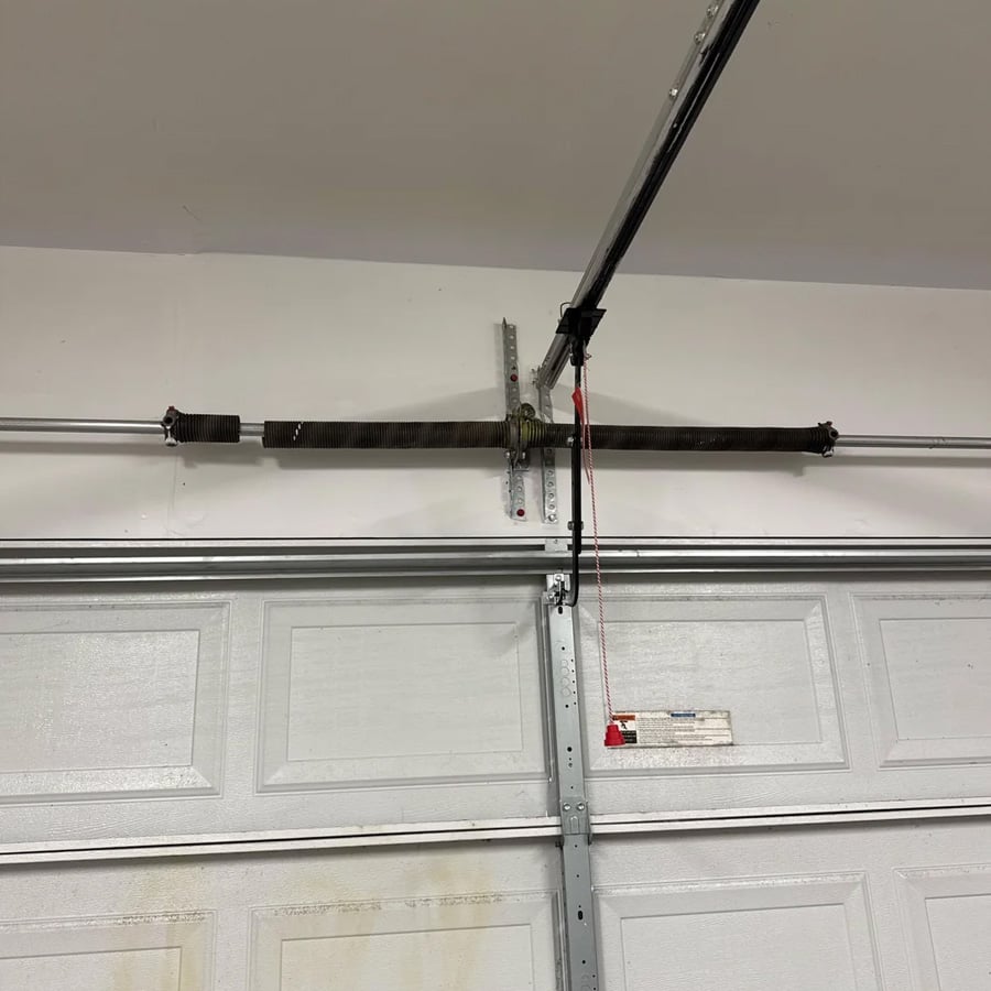A garage door torsion spring system is mounted above a closed white garage door. A red emergency release cord hangs down from the opener rail.