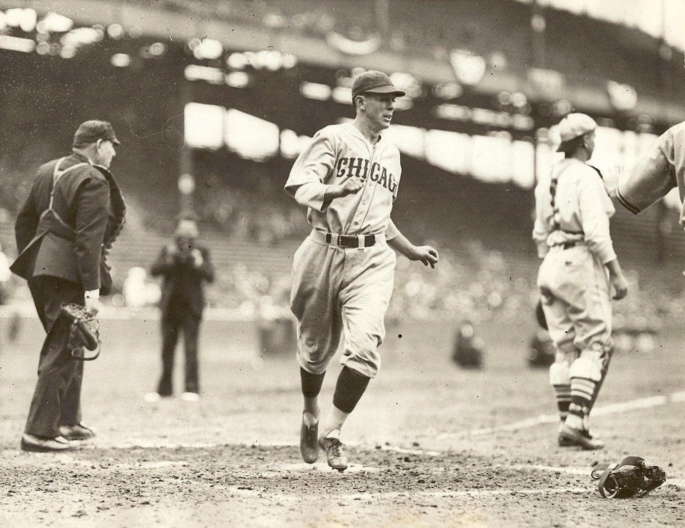 A baseball player in a “Chicago” uniform runs on a dirt field toward home plate, while other players and an umpire watch. The stadium seats and crowd are blurred in the background.